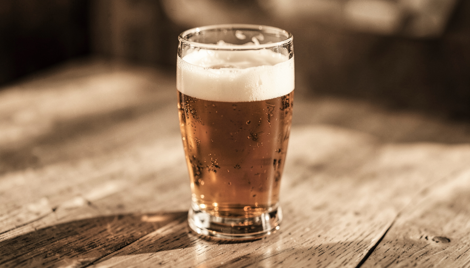 Close-up van een glas bier op een houten tafel, in warme beige en koperen tinten, zachte middaglicht.